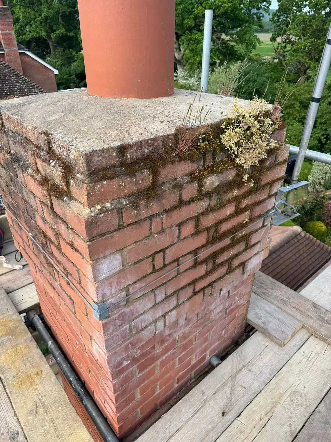 Brick chimney with moss and small plants growing on top, surrounded by wooden scaffolding.