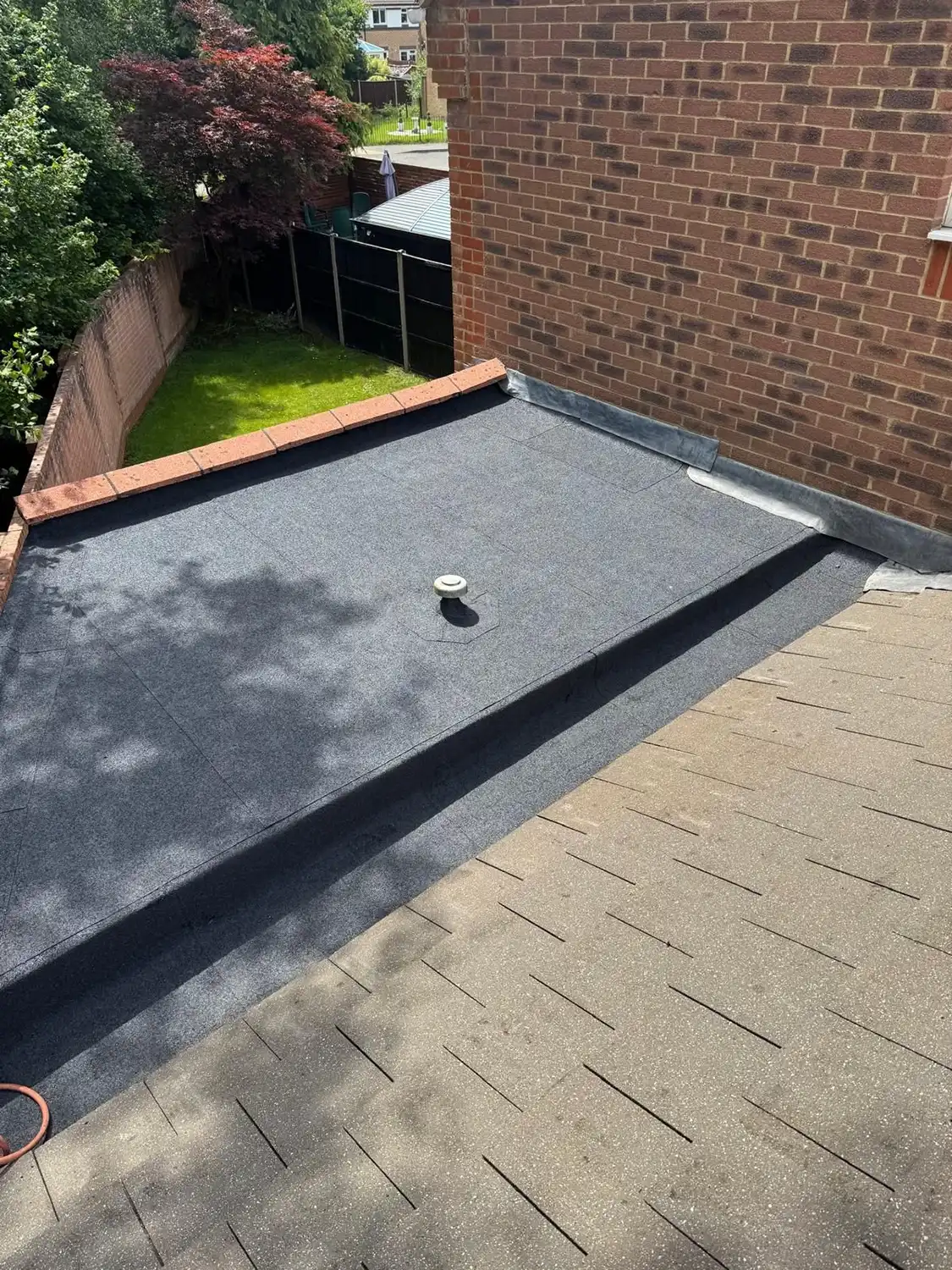 Flat roof with fresh grey waterproofing membrane adjacent to a brick wall and shingled sloped roof.