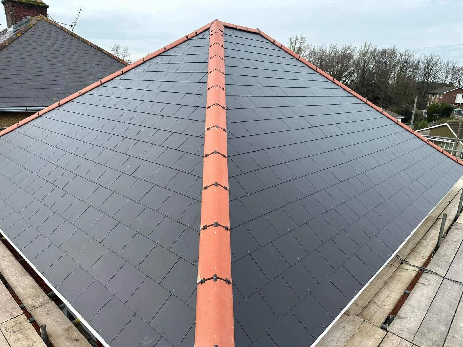 New black tiled roof with orange ridge tiles viewed from scaffolding at the roof edge.